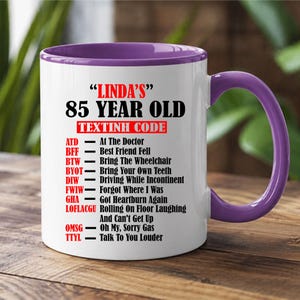 May include: A white ceramic mug with a purple interior and handle. The mug features the text "LINDA'S" 85 YEAR OLD TEXTINH CODE. The text includes abbreviations and their meanings, such as "ATD - At The Doctor" and "BFF - Best Friend Fell".