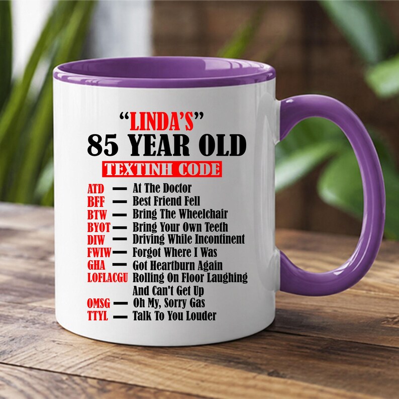 May include: A white ceramic mug with a purple interior and handle. The mug features the text "LINDA'S" 85 YEAR OLD TEXTINH CODE. The text includes abbreviations and their meanings, such as "ATD - At The Doctor" and "BFF - Best Friend Fell".