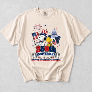 May include: Cream-colored t-shirt featuring Snoopy and Woodstock celebrating the 250th Anniversary of the United States of America. Snoopy wears a patriotic hat and holds a flag. The design includes the Capitol building, fireworks, and the text "250th Anniversary 1776-2026".