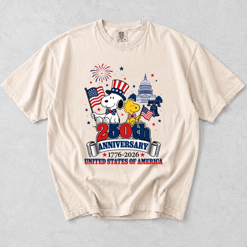 May include: Cream-colored t-shirt featuring Snoopy and Woodstock celebrating the 250th Anniversary of the United States of America. Snoopy wears a patriotic hat and holds a flag. The design includes the Capitol building, fireworks, and the text "250th Anniversary 1776-2026".
