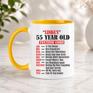 May include: A white ceramic mug with a yellow handle and interior. The mug features the text "LINDA'S 55 YEAR OLD TEXTINH CODE" with a list of humorous acronyms and their meanings, such as "ATD - At The Doctor."