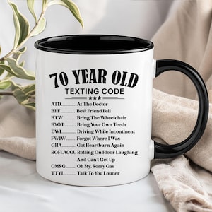 May include: A white ceramic mug with a black interior and handle. The mug features the text "70 YEAR OLD TEXTING CODE" in black, along with a list of texting acronyms and their meanings. A humorous gift idea.