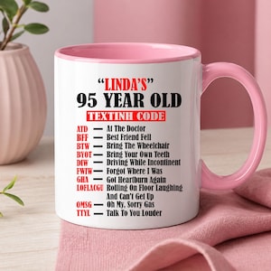 May include: White ceramic mug with a pink interior and handle. The mug features the text "LINDA'S" and "95 YEAR OLD TEXTINH CODE" in black, with a list of abbreviations and their meanings in red and black.