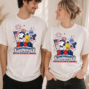 May include: White t-shirts featuring a patriotic design for the 250th anniversary of the United States. The design includes Snoopy and Woodstock, the American flag, the Capitol building, and the text "250th Anniversary 1776-2026 United States of America."