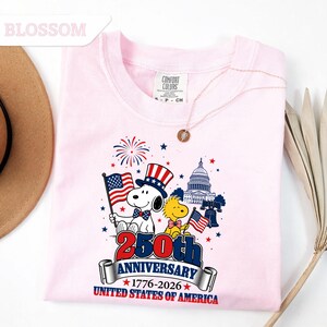 May include: A light pink sweatshirt featuring a graphic celebrating the 250th anniversary of the United States of America. The design includes Snoopy, Woodstock, the Capitol building, and the American flag. The text reads "250th Anniversary 1776-2026 United States of America."