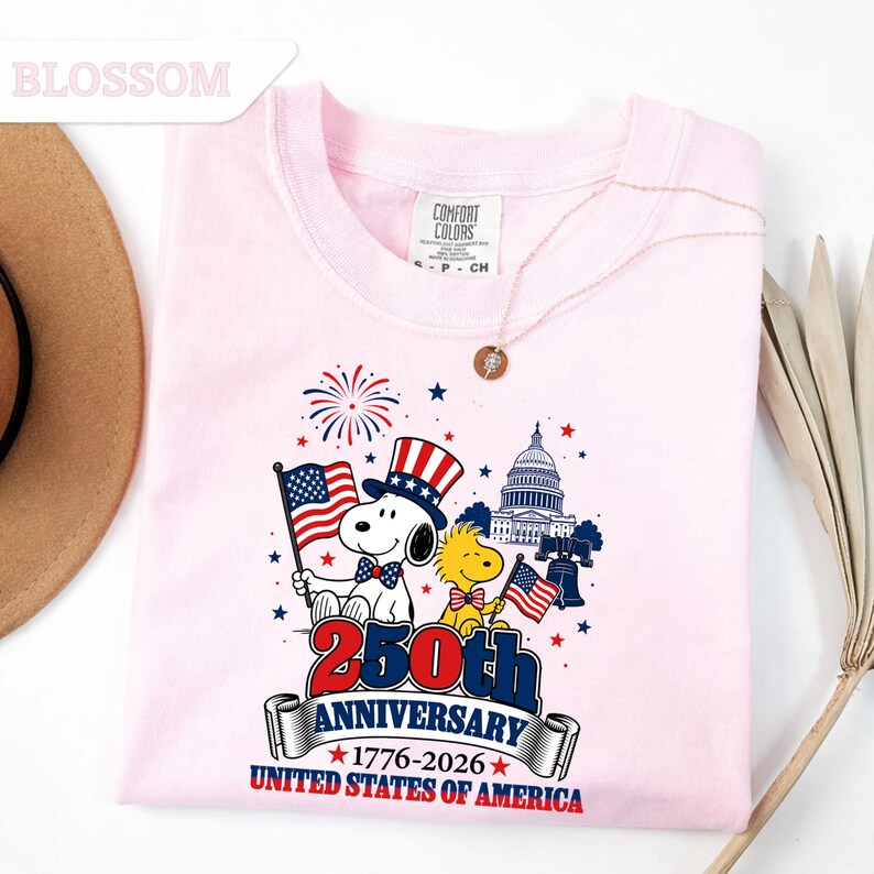 May include: A light pink sweatshirt featuring a graphic celebrating the 250th anniversary of the United States of America. The design includes Snoopy, Woodstock, the Capitol building, and the American flag. The text reads "250th Anniversary 1776-2026 United States of America."