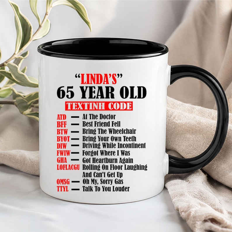 May include: A white ceramic mug with a black interior and handle. The mug displays the text "LINDA'S" and "65 YEAR OLD" in black, with a red banner reading "TEXTINH CODE". Below are humorous abbreviations and their meanings.