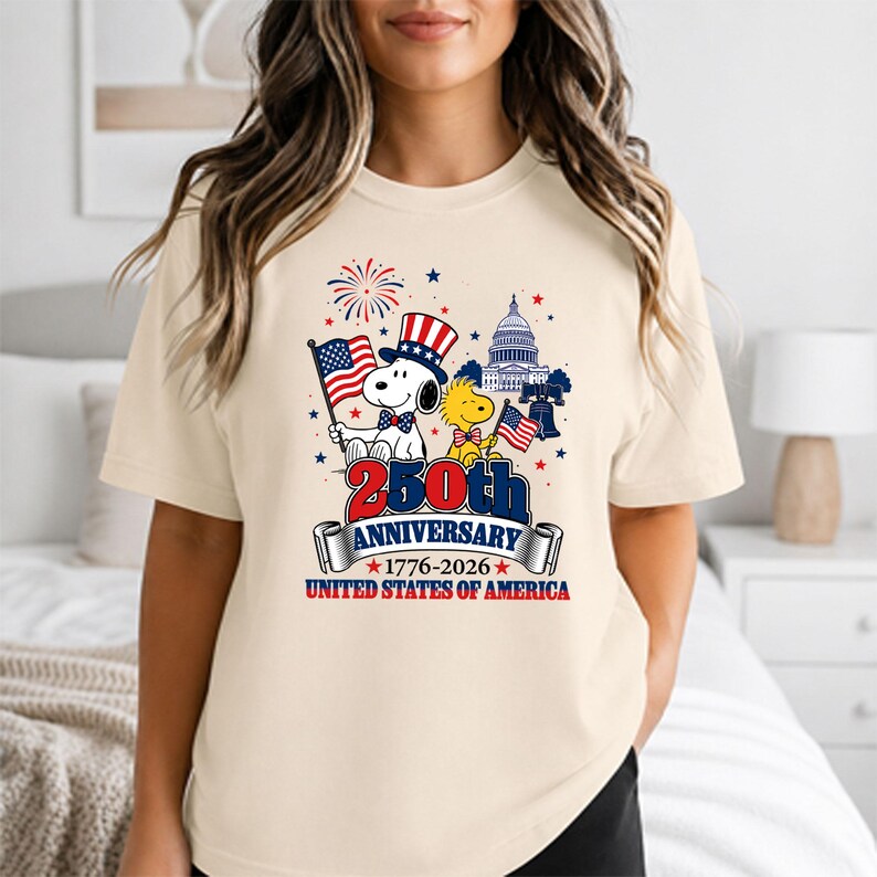 May include: A cream-colored t-shirt featuring a patriotic design for the 250th anniversary of the United States. The graphic includes Snoopy in a patriotic hat, Woodstock, the Capitol building, and the text "250th Anniversary 1776-2026 United States of America."