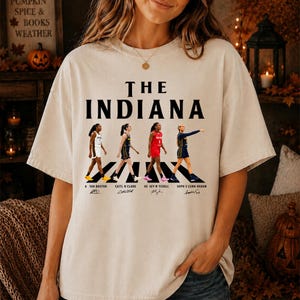 May include: A beige short-sleeve t-shirt featuring the text THE INDIANA in black lettering above a graphic of four basketball players walking on a crosswalk. Below each player are their names and signatures: Aaliyah Boston, Caitlin Clark, Kelsey Mitchell, and Sophie Cunningham.