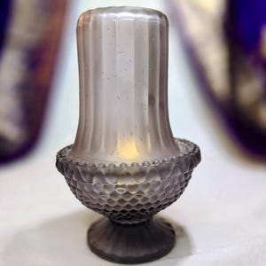 Puede incluir: Un portavelas de cristal vintage, en tonos grises. El portavelas tiene una base texturizada, similar a una uña, y una parte superior cilíndrica acanalada. La base está soportada por un pedestal corto y redondo. El diseño general es elegante y antiguo.