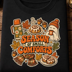 Może przedstawiać: Czarna bluza z grafiką i napisem "SEASON OF SMALL COMFORTS" w kolorze pomarańczowym. Wzór przedstawia termos, bułeczki cynamonowe, duszka w czapce, żołędzie, liście klonu oraz szalik w kratę w jesiennych barwach.