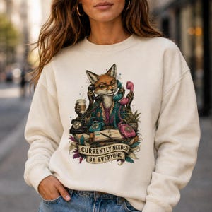 Może przedstawiać: Kremowa bluza z okrągłym dekoltem z grafiką przedstawiającą lisa w okularach, otoczonego wieloma telefonami w stylu vintage, filiżanką kawy i otwartym terminarzem, z napisem "CURRENTLY NEEDED BY EVERYONE" na banerze.