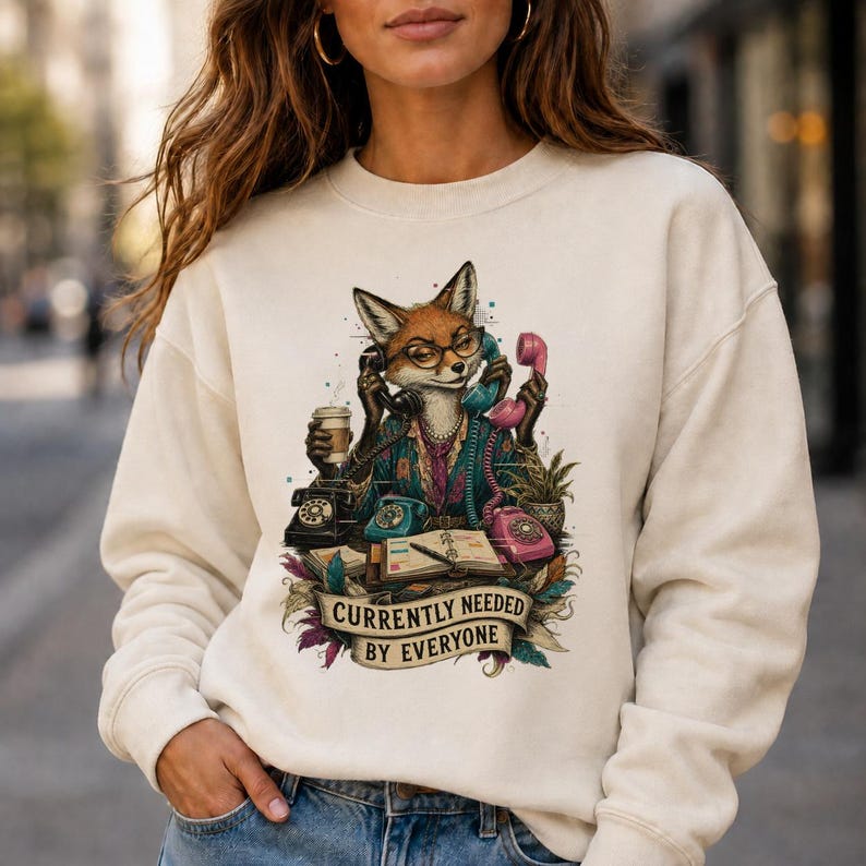 Może przedstawiać: Kremowa bluza z okrągłym dekoltem z grafiką przedstawiającą lisa w okularach, otoczonego wieloma telefonami w stylu vintage, filiżanką kawy i otwartym terminarzem, z napisem "CURRENTLY NEEDED BY EVERYONE" na banerze.