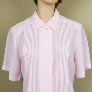 May include: A pink, short-sleeved blouse with a collared neckline and a row of vertical pleats down the front. The blouse has a button-up closure and a pearl necklace.