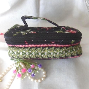 May include: A rectangular quilted fabric case with a top handle and a zipper closure. The upper section features a black floral pattern, while the lower section displays a green and white leaf print with a pink trim accenting the middle.
