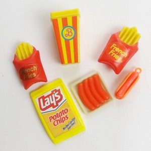 May include: A set of miniature plastic food items, including a yellow and red french fries container, a yellow and red bag of french fries, a yellow bag of Lay's potato chips, a white tray with four red hot dogs, and a red hot dog.