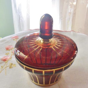 May include: A red glass jar with a gold lid and a gold knob. The jar has gold stripes around the bottom and a gold sunburst design on the lid.