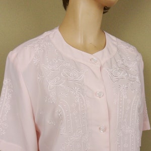 May include: A pale pink button-up blouse with short sleeves and white embroidered floral designs. The blouse has a mandarin collar and a row of white buttons down the front.