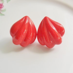 May include: Two bright red, sculptural earrings with a swirling, petal-like design. The earrings are set on a white, floral-decorated plate, creating a striking contrast. The earrings have a glossy finish and a unique, organic shape.