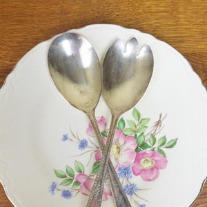 May include: A pair of silver salad servers with ornate handles, resting on a white porcelain plate with a floral design featuring pink roses and blue flowers.