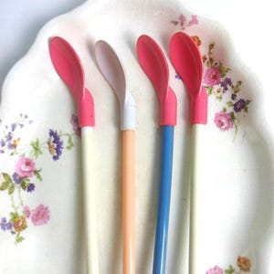 May include: Four long-handled spoons with colorful plastic heads and slender stems. The spoons feature pink, white, and blue heads attached to cream, peach, and blue handles. These utensils are arranged vertically, showcasing their distinct color combinations.