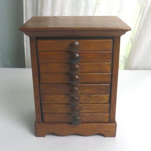 May include: A small, rectangular wooden chest featuring seven stacked drawers. Each drawer is equipped with a single, centered, round wooden knob. The chest has a flat top with a slight overhang and a scalloped base, finished in a medium brown wood tone.