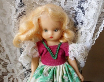 Puede incluir: Una muñeca de porcelana vintage con cabello rubio, vestida con un vestido verde y blanco con un corpiño rosa y un collar de cuentas verdes. La muñeca está posada sobre un fondo de encaje blanco.