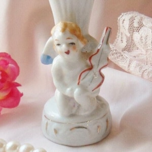 May include: A white porcelain vase with a gold trim and a cherub figurine holding a violin. The vase is decorated with gold accents and has a delicate design.