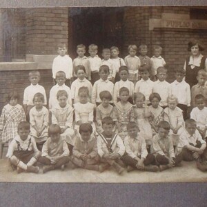 May include: A black and white photo of a class of children standing in front of a brick building. The children are all wearing clothing from the early 1900s. The building has the words "Public School" above the entrance.