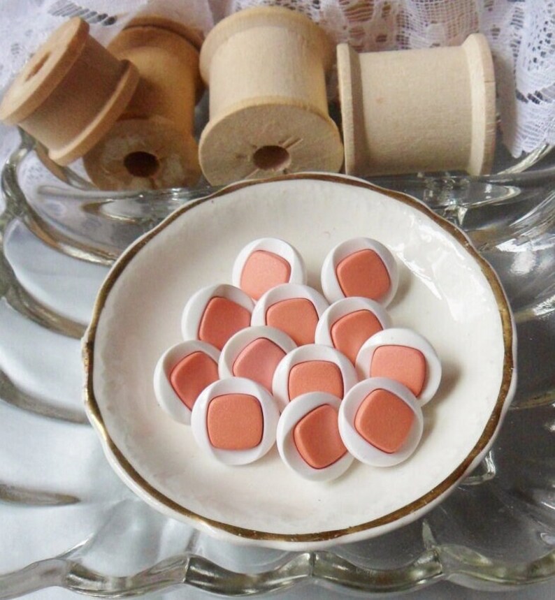 Puede incluir: Una colecci&oacute;n de 10 botones de color blanco y melocot&oacute;n con una forma cuadrada en el centro. Los botones est&aacute;n dispuestos en c&iacute;rculo sobre un plato blanco.