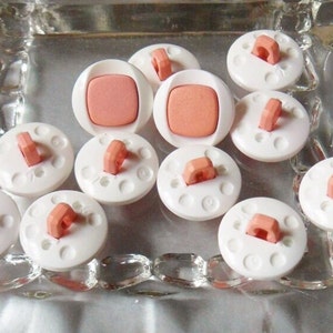 Puede incluir: Una colecci&oacute;n de diez botones de pl&aacute;stico blanco con un centro rosa y un lazo en la parte superior. Los botones est&aacute;n dispuestos en un plato de vidrio.