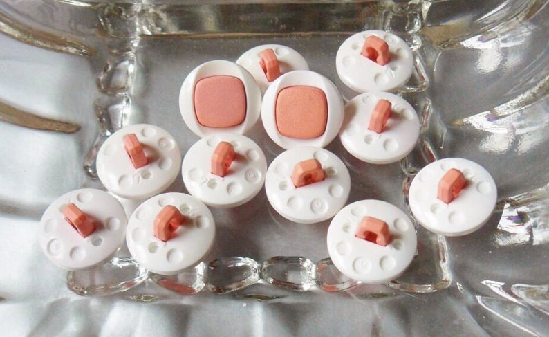 Puede incluir: Una colecci&oacute;n de diez botones de pl&aacute;stico blanco con un centro rosa y un lazo en la parte superior. Los botones est&aacute;n dispuestos en un plato de vidrio.