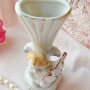 May include: A white porcelain vase with a gold trim and a figurine of a child holding a red and white toy. The vase is decorated with a blue and white floral pattern.