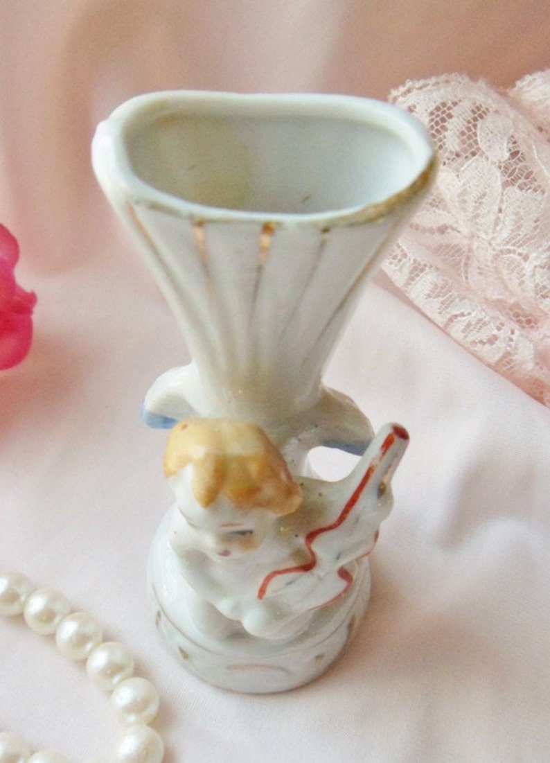 May include: A white porcelain vase with a gold trim and a figurine of a child holding a red and white toy. The vase is decorated with a blue and white floral pattern.