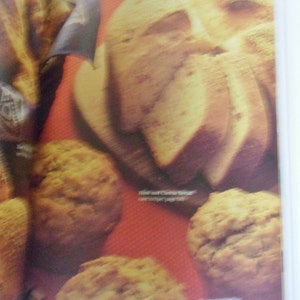 May include: A close-up of a cookbook page featuring a variety of baked goods, including a loaf of bread shaped like a skull, muffins, and a loaf of bread. The text on the page reads "Ham and Cheese Bread (see recipe page 58)" and "Mammoth Muffins (see recipe page 50)."