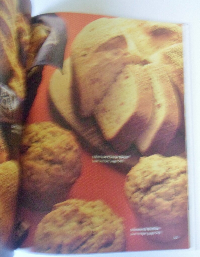 May include: A close-up of a cookbook page featuring a variety of baked goods, including a loaf of bread shaped like a skull, muffins, and a loaf of bread. The text on the page reads "Ham and Cheese Bread (see recipe page 58)" and "Mammoth Muffins (see recipe page 50)."