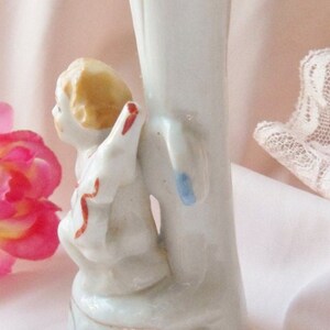 May include: A white porcelain vase with a gold trim features a small angel figurine with a red and white ribbon. The angel is sitting on a ledge with its wings spread out.