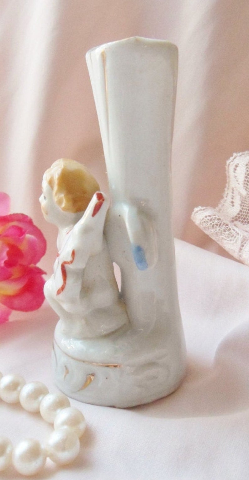 May include: A white porcelain vase with a gold trim features a small angel figurine with a red and white ribbon. The angel is sitting on a ledge with its wings spread out.