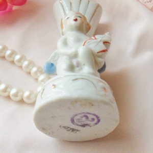 May include: White porcelain figurine of an angel holding a small white bird. The angel is sitting on a round base with gold accents. The base has a purple stamp with the number "23" inside a circle.