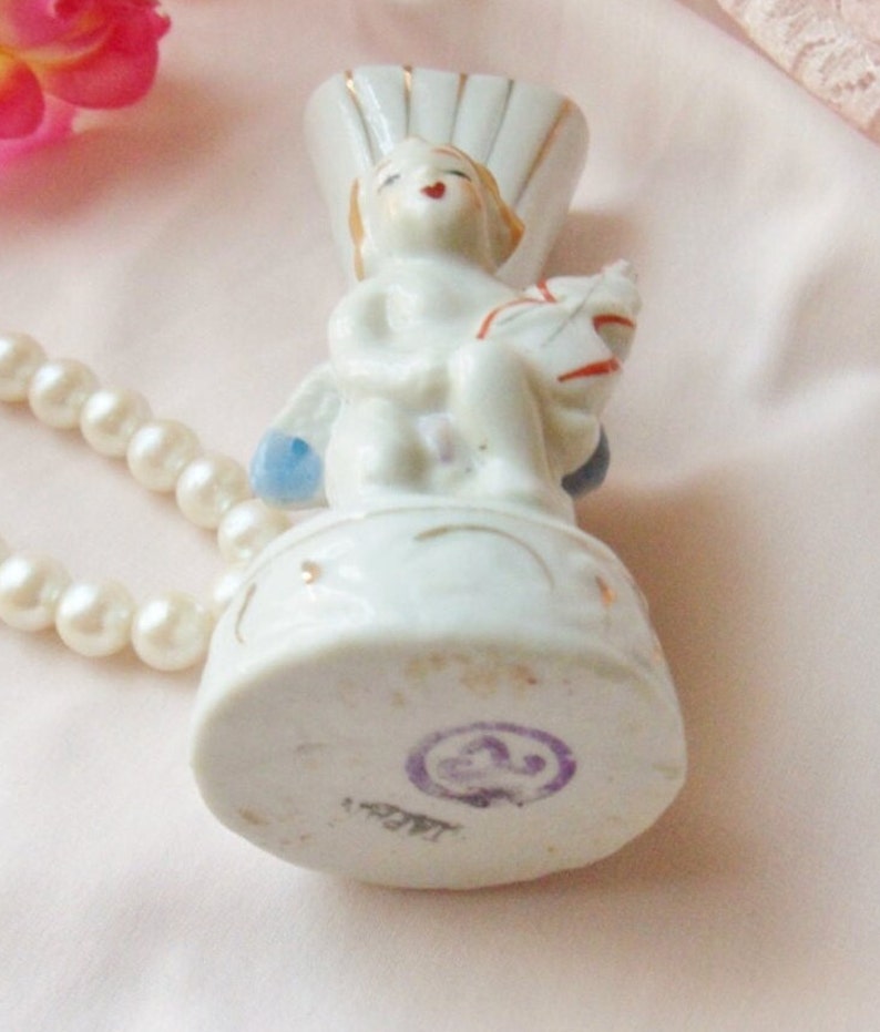 May include: White porcelain figurine of an angel holding a small white bird. The angel is sitting on a round base with gold accents. The base has a purple stamp with the number "23" inside a circle.