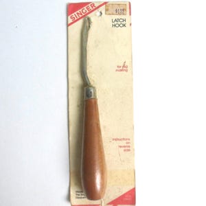 May include: A vintage Singer latch hook tool with a smooth wooden handle and a metal hook, mounted on its original rectangular cardboard packaging. The packaging features red and white branding, text reading LATCH HOOK, and instructions on reverse side.