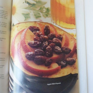 May include: A close-up of a halved acorn squash filled with sliced apples and raisins. The squash is on a dark brown surface and the text "Apple-Filled Squash" is printed below it.