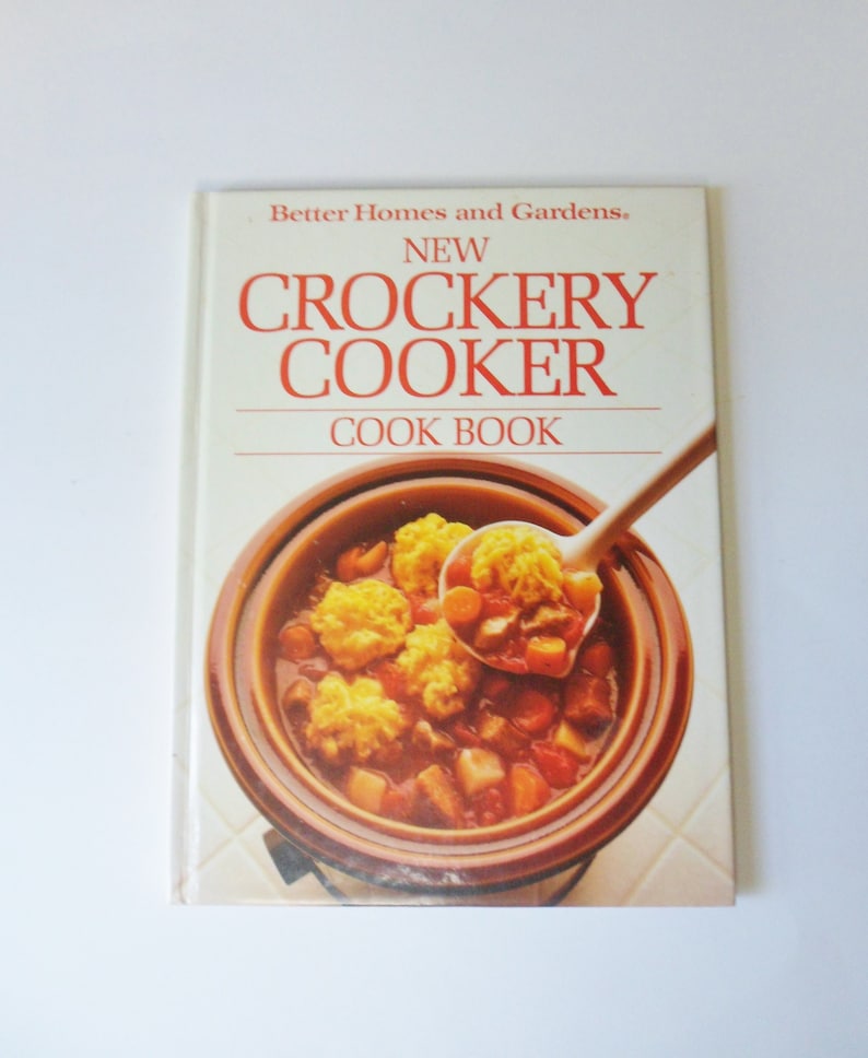 May include: A white cookbook with the title "New Crockery Cooker Cookbook" by Better Homes and Gardens. The cover features a close-up of a crock pot filled with a stew with dumplings.