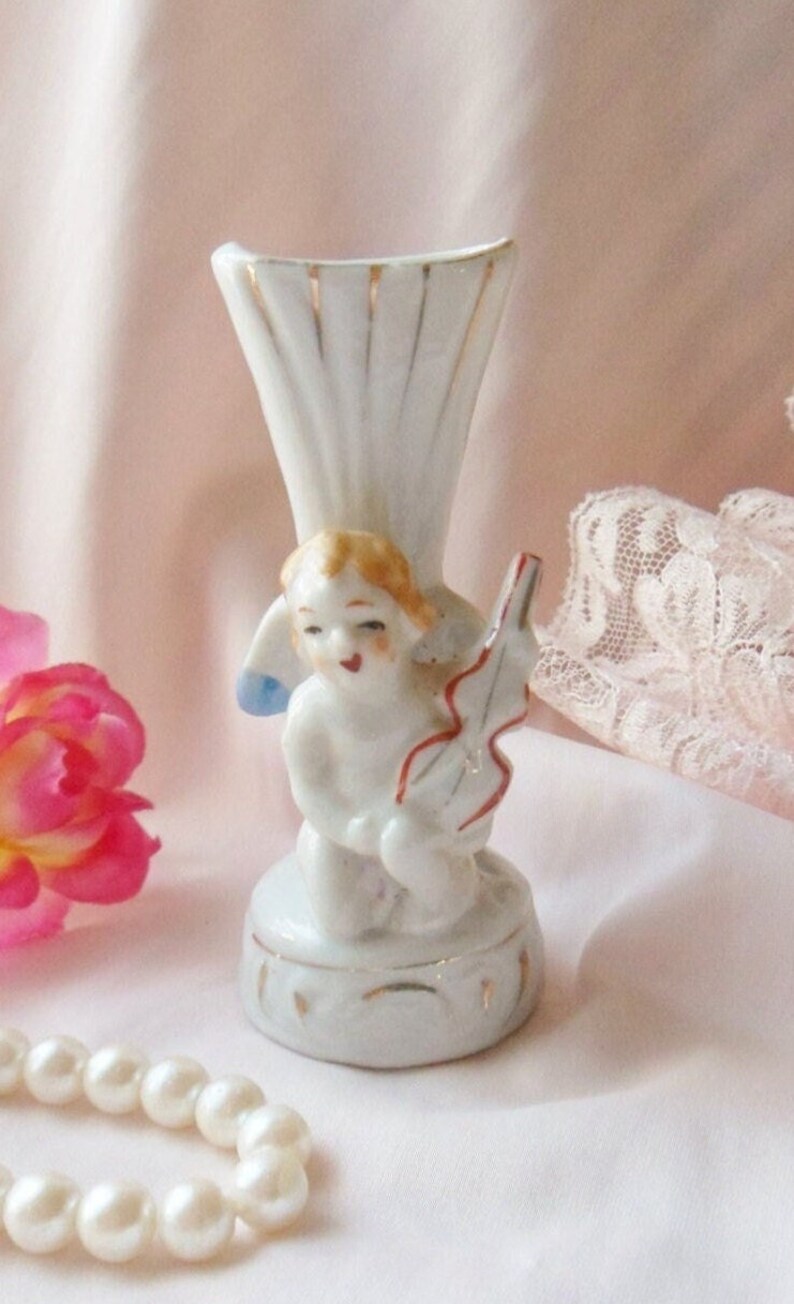 May include: A white porcelain vase with a gold trim and a cherub figurine holding a violin. The vase is decorated with gold accents and has a delicate design.