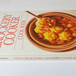 May include: A white cookbook with red text titled "New Crockery Cooker Cook Book" by Better Homes and Gardens. The cover features a close-up of a brown crock pot filled with a stew and dumplings.