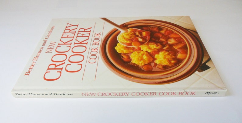 May include: A white cookbook with red text titled "New Crockery Cooker Cook Book" by Better Homes and Gardens. The cover features a close-up of a brown crock pot filled with a stew and dumplings.