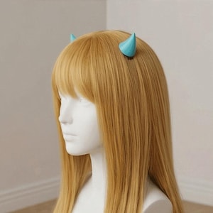 Puede incluir: Una cabeza de maniquí con cabello largo y rubio claro y flequillo, peinada con dos pequeños cuernos de diablo azul claro. Los cuernos están colocados en la parte superior de la cabeza, agregando un toque caprichoso al peinado.