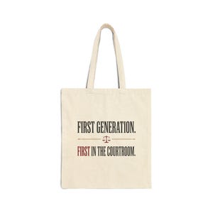 May include: A cream-coloured tote bag featuring the text "FIRST GENERATION." and "FIRST IN THE COURTROOM." in black, with a small scale graphic between the phrases. The bag has two handles and is made of a canvas-like material.