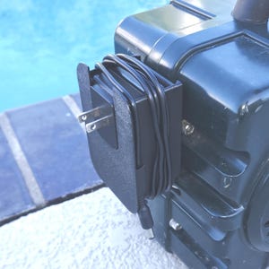 May include: A black power adapter with a two-prong plug is attached to a dark green device. The adapter's cord is neatly wrapped around the device. The background features a blue water surface.