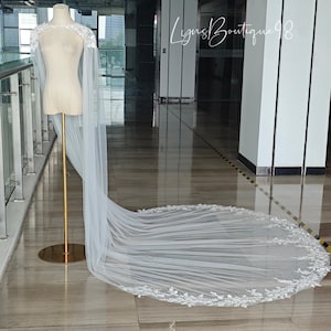 May include: A white bridal cape and veil displayed on a mannequin. The cape features delicate lace detailing around the shoulders, and the veil extends into a long train with matching lace trim. The mannequin is on a gold stand.